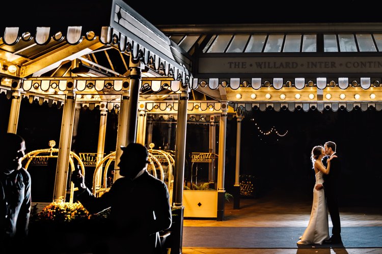 A bride and groom sharing a dance at night near a brightly lit amusement park ride, with silhouettes of two people talking in the foreground.