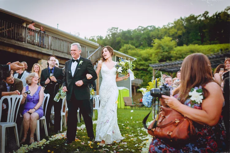 Bride walking down the aisle with her father during a wedding ceremony, surrounded by seated guests and trees in the background.