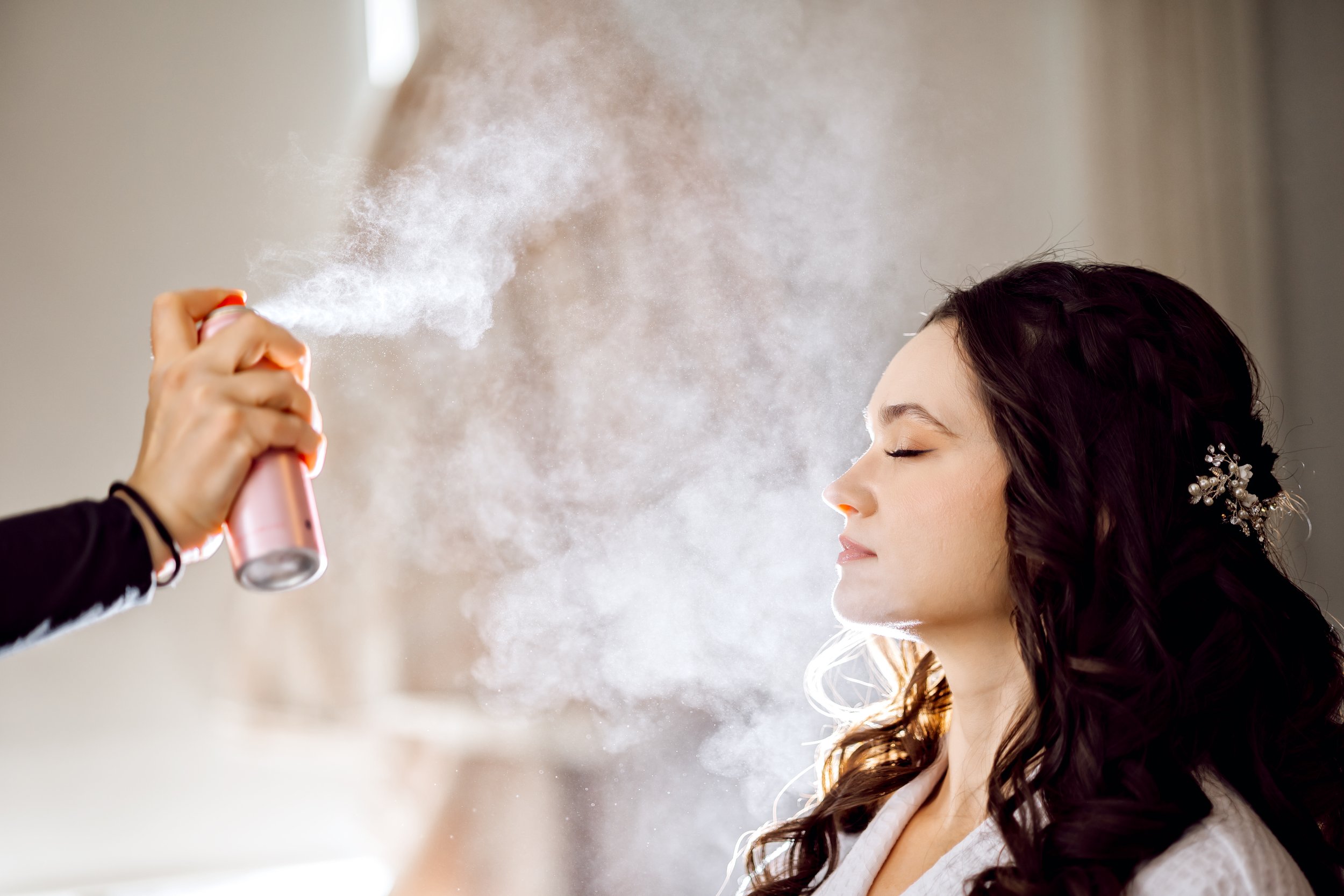A woman with dark, curled hair and a decorative hair accessory has her eyes closed as someone sprays her with a can of mist or spray, creating a cloud of vapor around her face.
