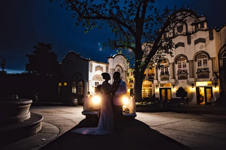 A silhouette of a couple standing in front of a vintage car at night outside a large illuminated mansion, with trees and a dark sky in the background.