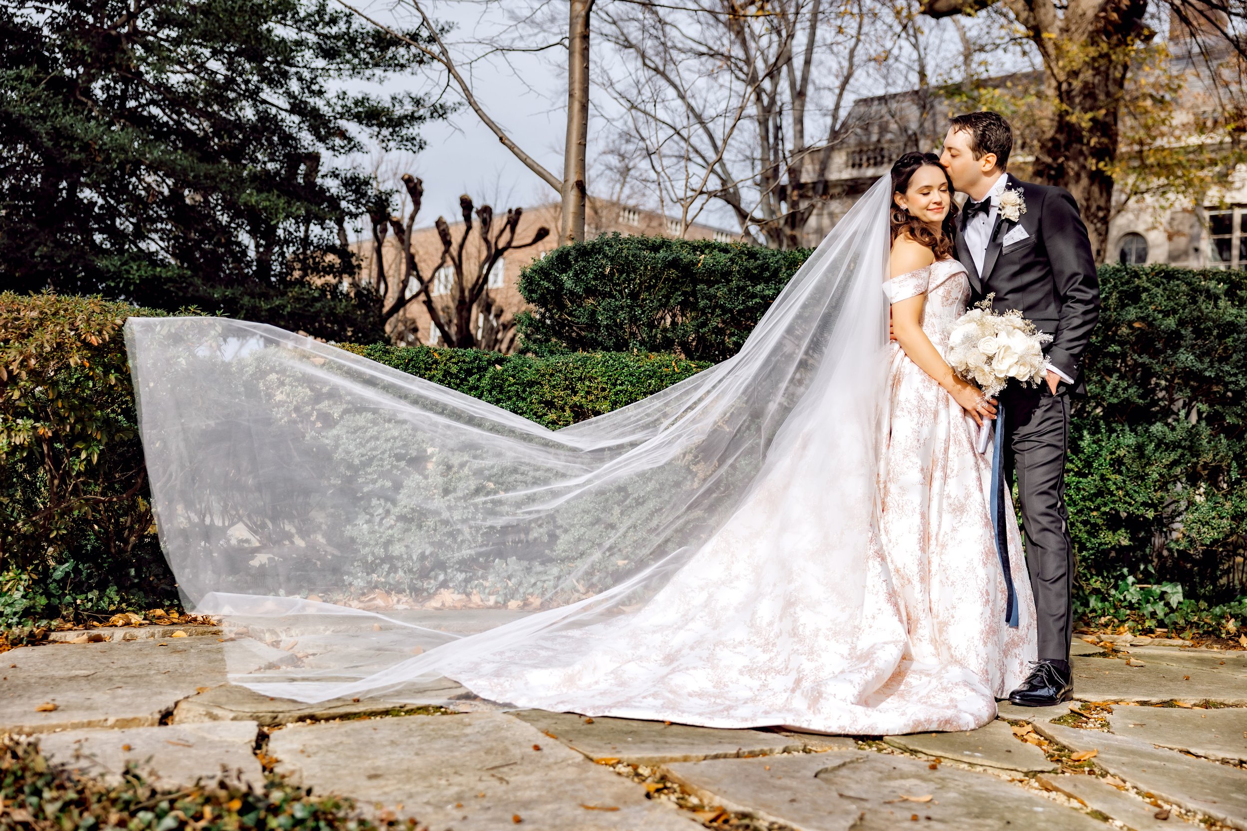 A bride and groom standing outdoors on a stone pathway, with the bride holding a bouquet and wearing a long veil that flows to the ground, while the groom kisses her forehead. They are surrounded by bushes and trees, with a building in the background