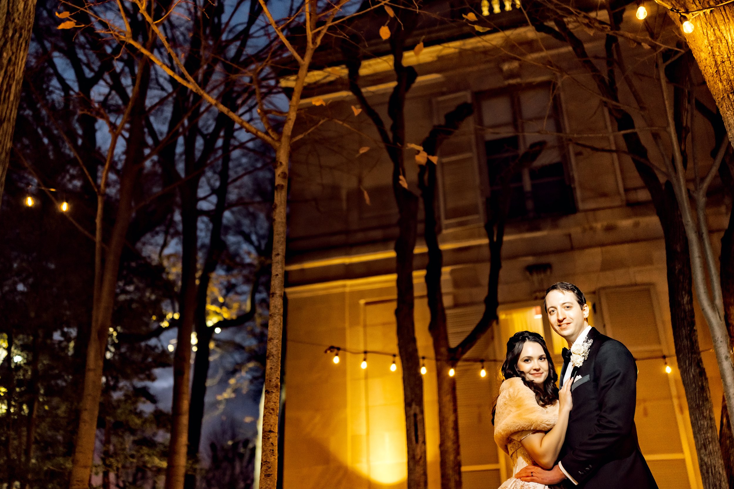 A newlywed couple dancing outdoors at night, surrounded by leafless trees and string lights, with a wooden house in the background.