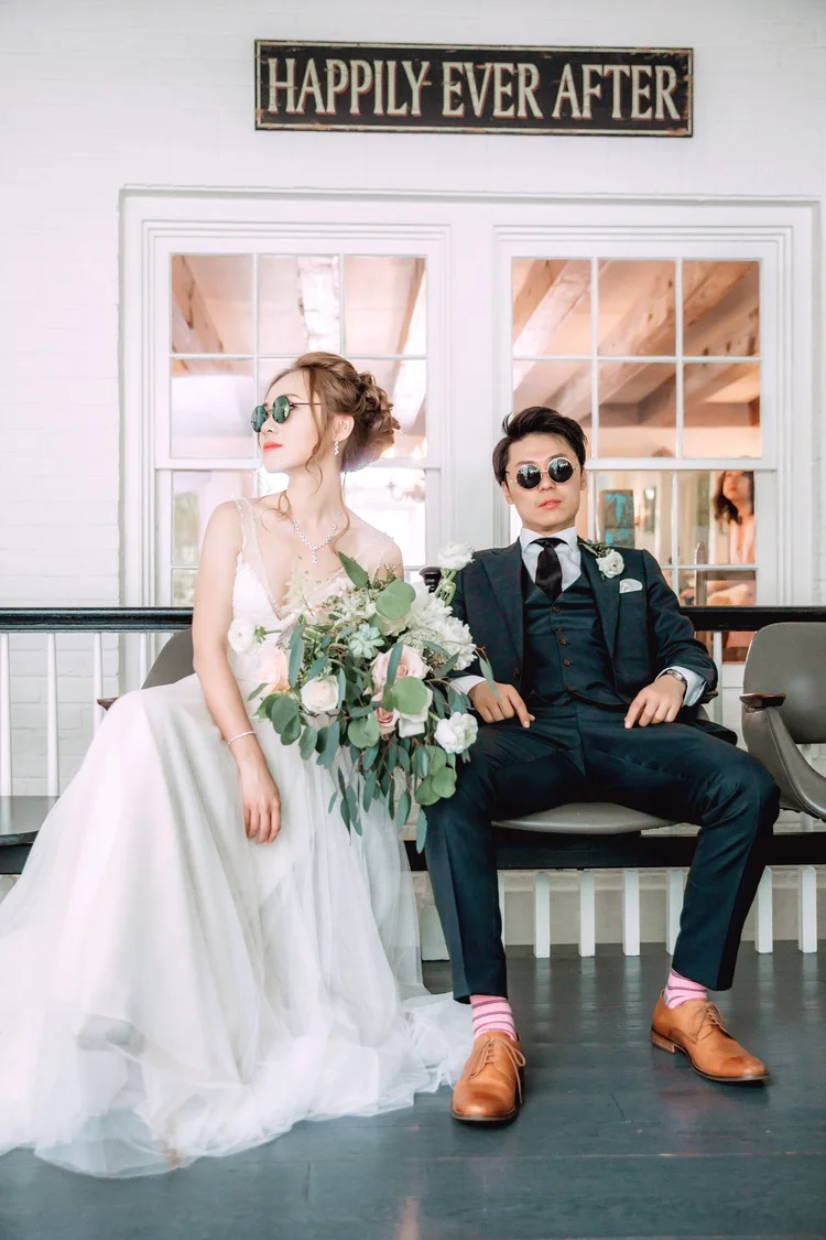 A bride and groom sitting on a bench in a wedding photo, with the bride holding a large bouquet of flowers and both wearing sunglasses. The bride is dressed in a white gown, and the groom is in a dark suit with pink striped socks. They are indoors, w