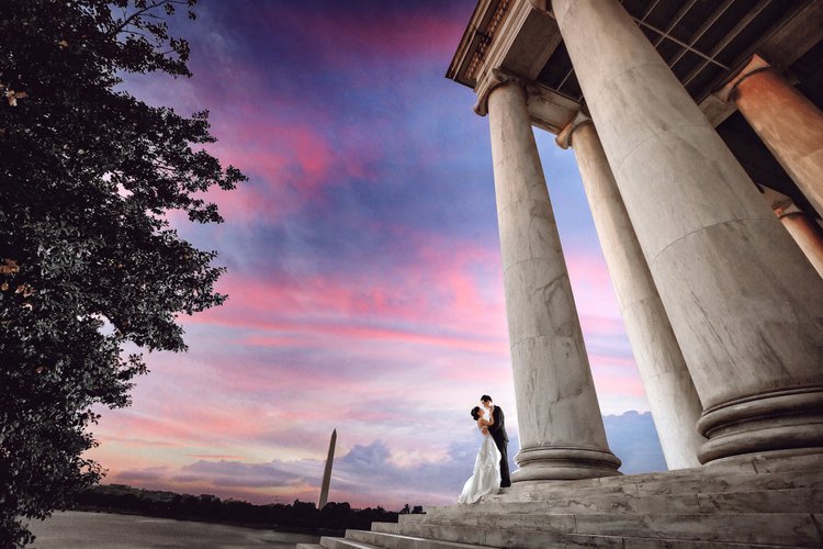 A bride and groom in wedding attire standing on steps, embracing in front of large marble columns, with the Washington Monument and a colorful sunset sky in the background.