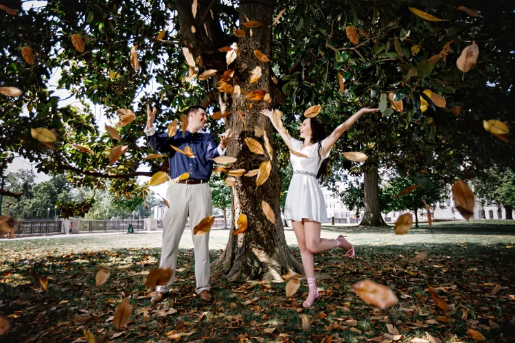 Two young adults, a man and a woman, playing and having fun under a large tree in a park, with leaves falling around them.