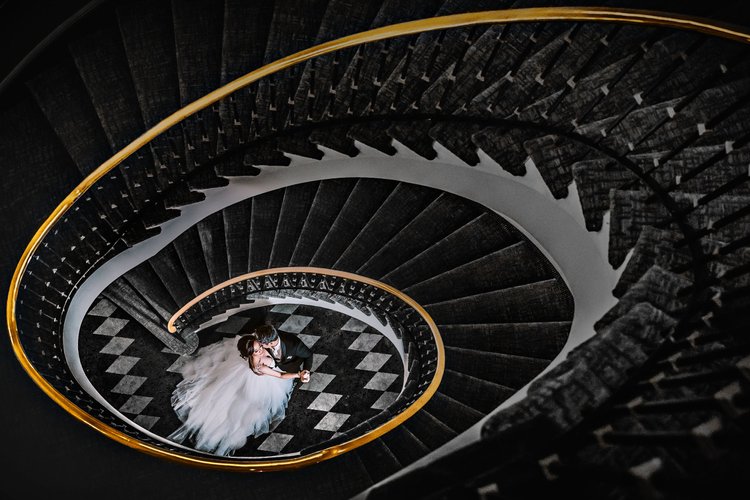 An overhead view of a bride and groom dancing on a black and white checkered floor at the bottom of a spiral staircase with black steps and gold handrails.