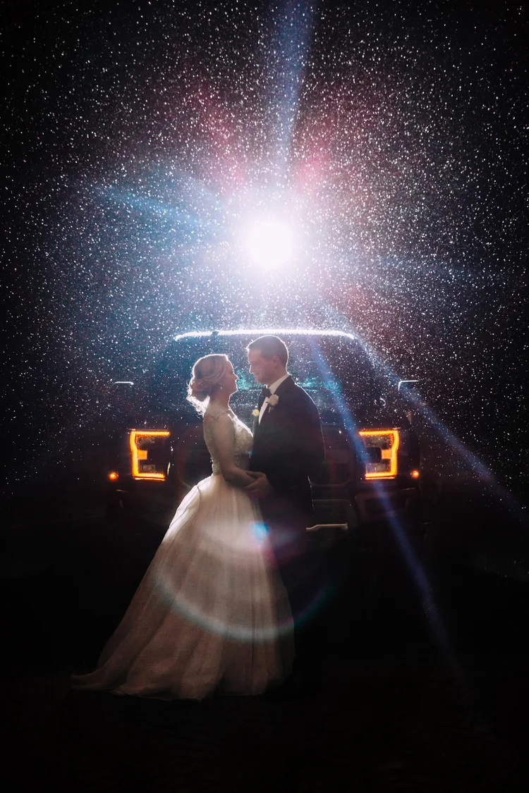 A bride and groom standing close together, holding hands, silhouetted in front of a starry night sky with a bright light shining behind them and a vehicle with lit tail lights in the background.