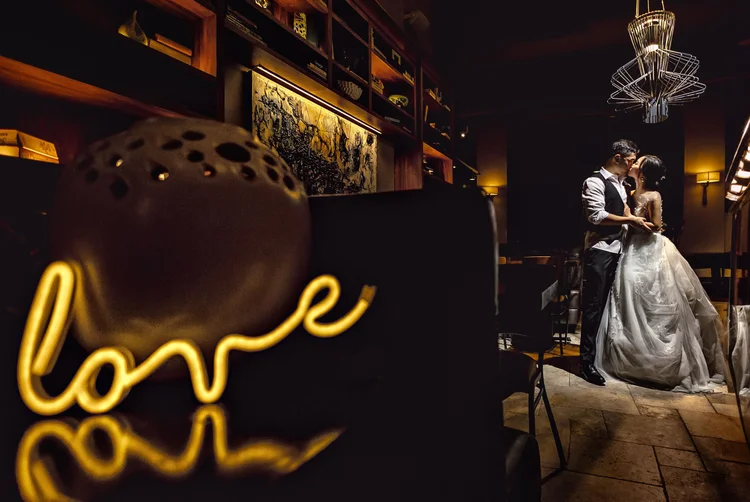 A couple on their wedding day sharing a kiss in a dimly lit restaurant or lounge. The bride wears a white wedding gown, and the groom is dressed in a vest with a shirt. In the foreground, there is a blurred decorative object shaped like a rounded, li