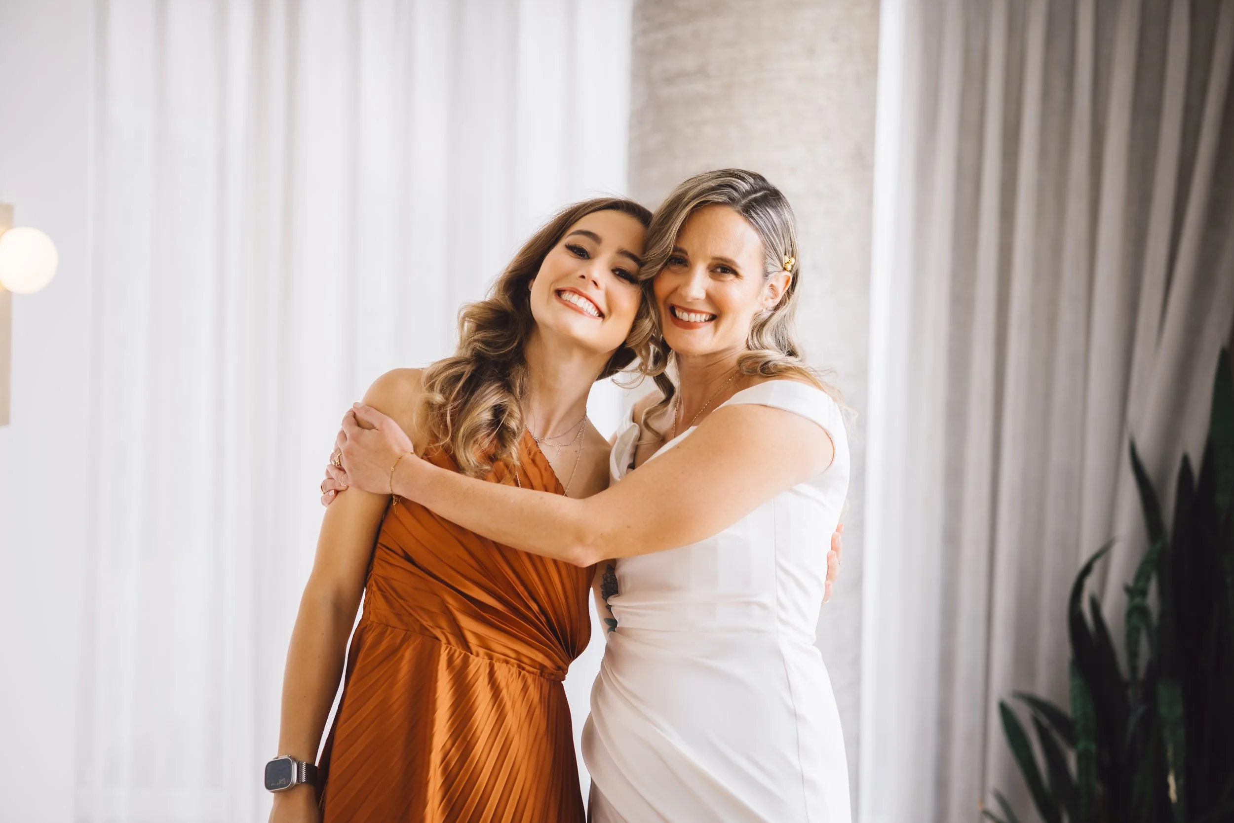 Two women smiling and hugging indoors, one in a white dress and the other in an orange dress.