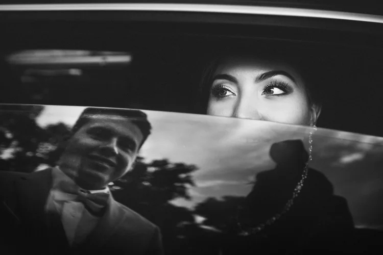 Black and white photo of a woman looking up, with her face partially obscured by a reflective surface showing a man in a tuxedo and bow tie smiling.