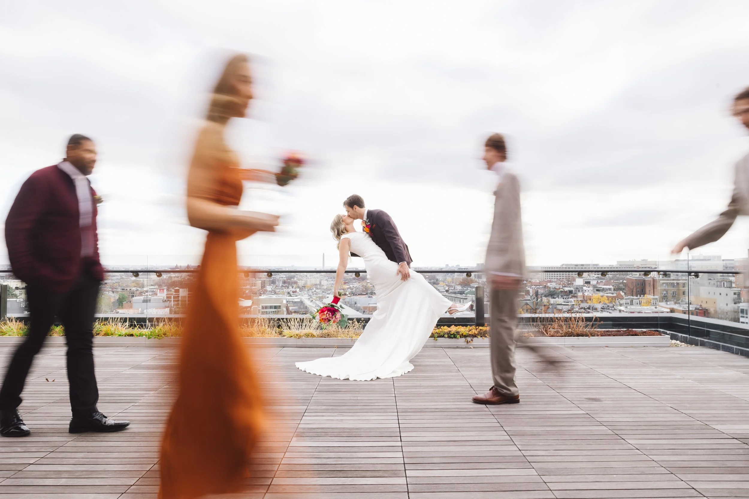A wedding scene with a bride and groom in the center, with the groom lifting the bride in a dip, all while the bride holds a bouquet of flowers. Four blurred guests are walking by in the foreground, on a rooftop with a cityscape in the background on 