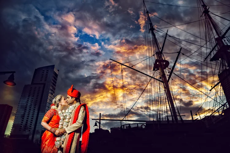 A couple dressed in traditional Indian wedding attire sharing a kiss at sunset near a harbor, with a large sailing ship and city buildings in the background.