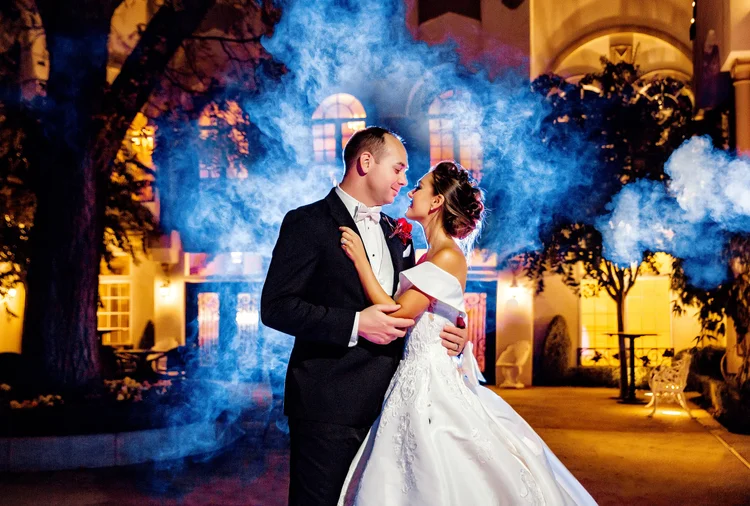 A bride and groom sharing a romantic dance at night outside a large, elegant building with warm lighting and trees with blue smoke in the background.
