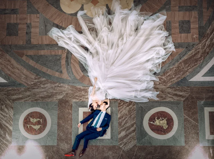 A newlywed couple lies on the patterned marble floor, with the bride in a voluminous white wedding gown and the groom in a dark tuxedo. They are facing each other, sharing an intimate kiss.