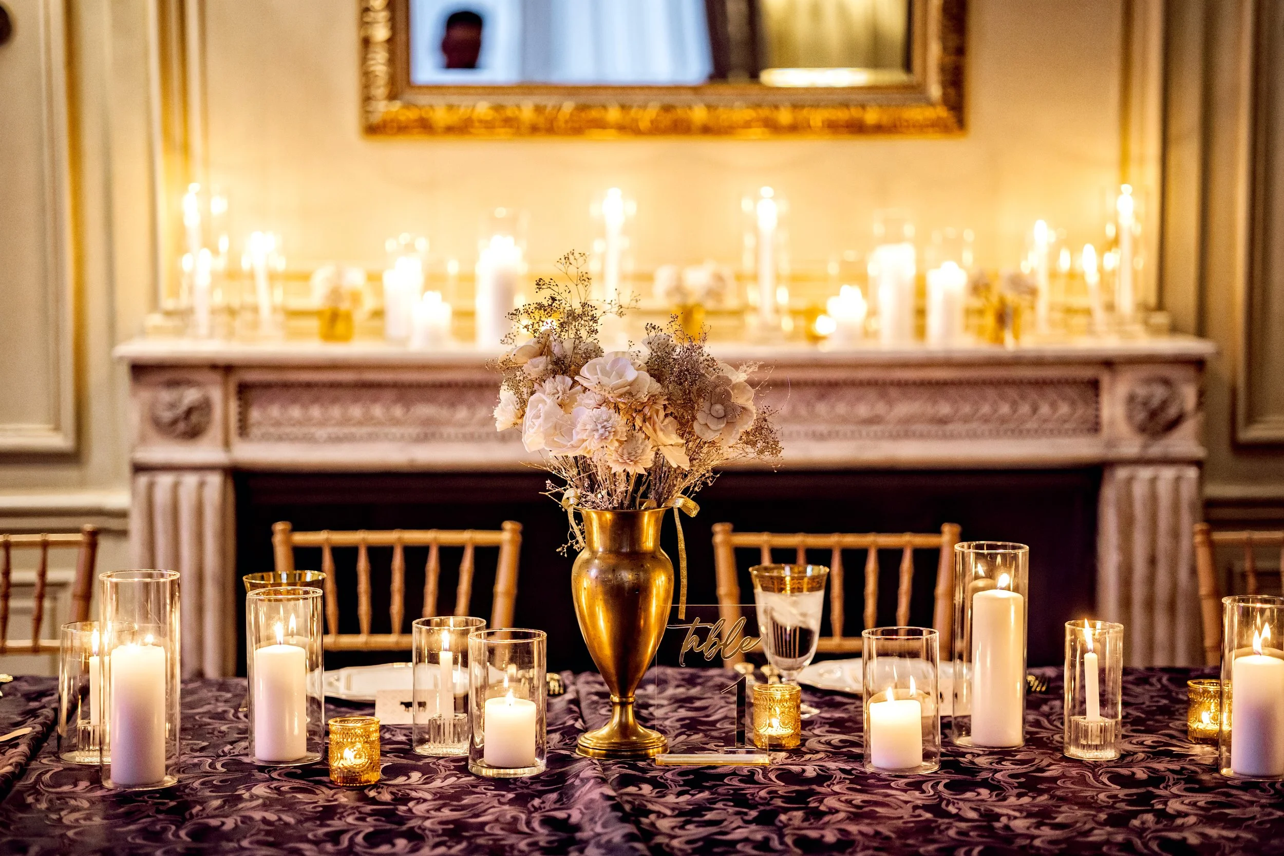 Elegant dining table decorated with white flowers in a gold vase, surrounded by lit candles in glass holders, with a fireplace and mirror in the background.