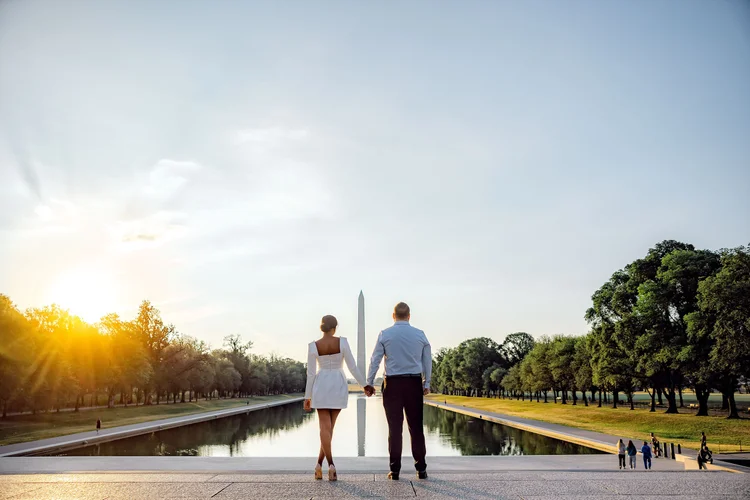 A couple holding hands walking towards the Washington Monument in Washington, D.C., during sunset, near the Reflecting Pool surrounded by trees.