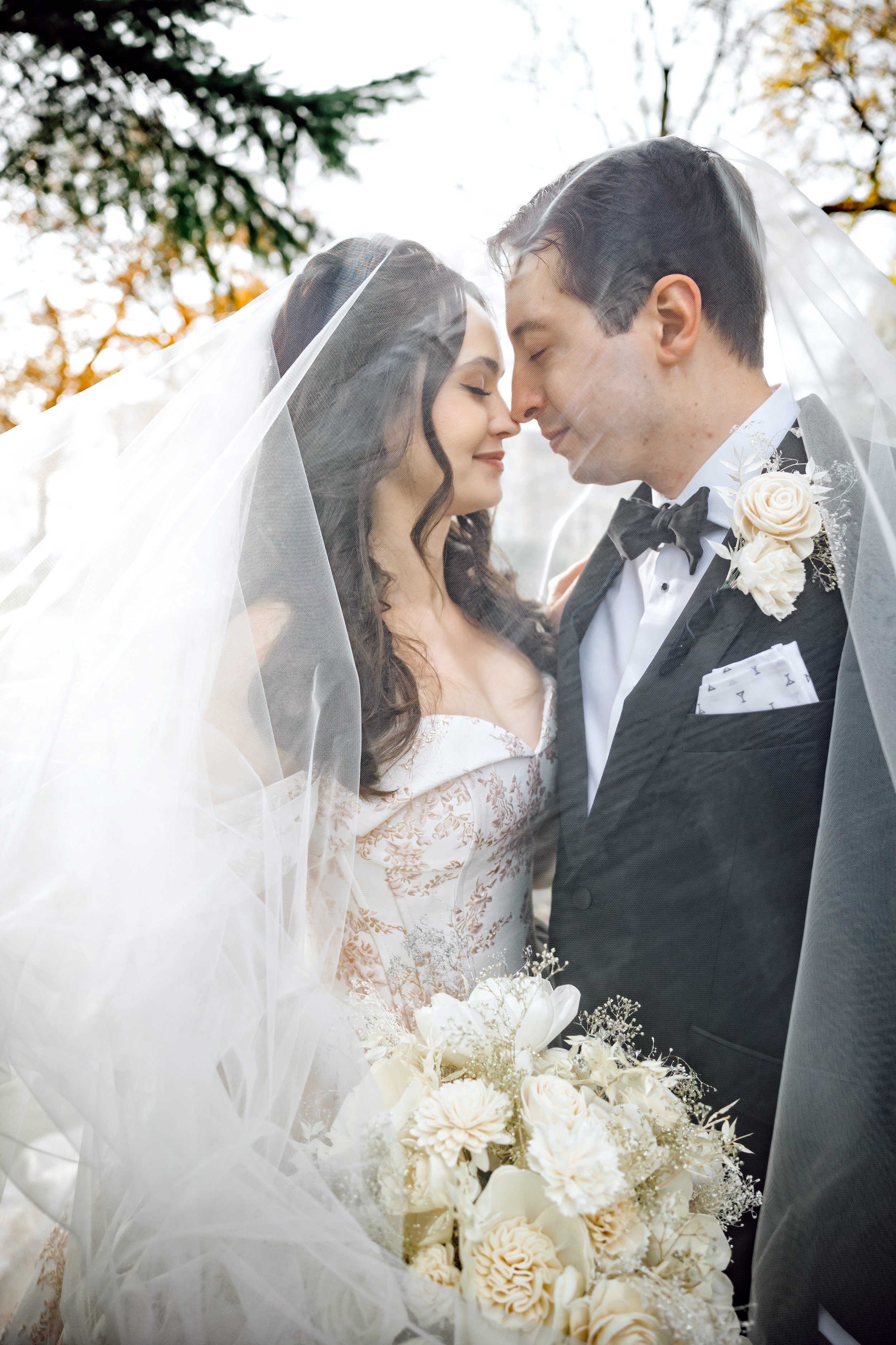 A bride and groom sharing a kiss, with their foreheads touching and eyes closed, under a wedding veil outdoors. The bride holds a bouquet of white flowers, and they are dressed in formal wedding attire, with the groom wearing a tuxedo and bow tie, an
