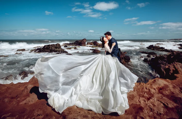 A bride and groom in wedding attire share a kiss on rocky shore by the ocean with waves and a blue sky in the background.