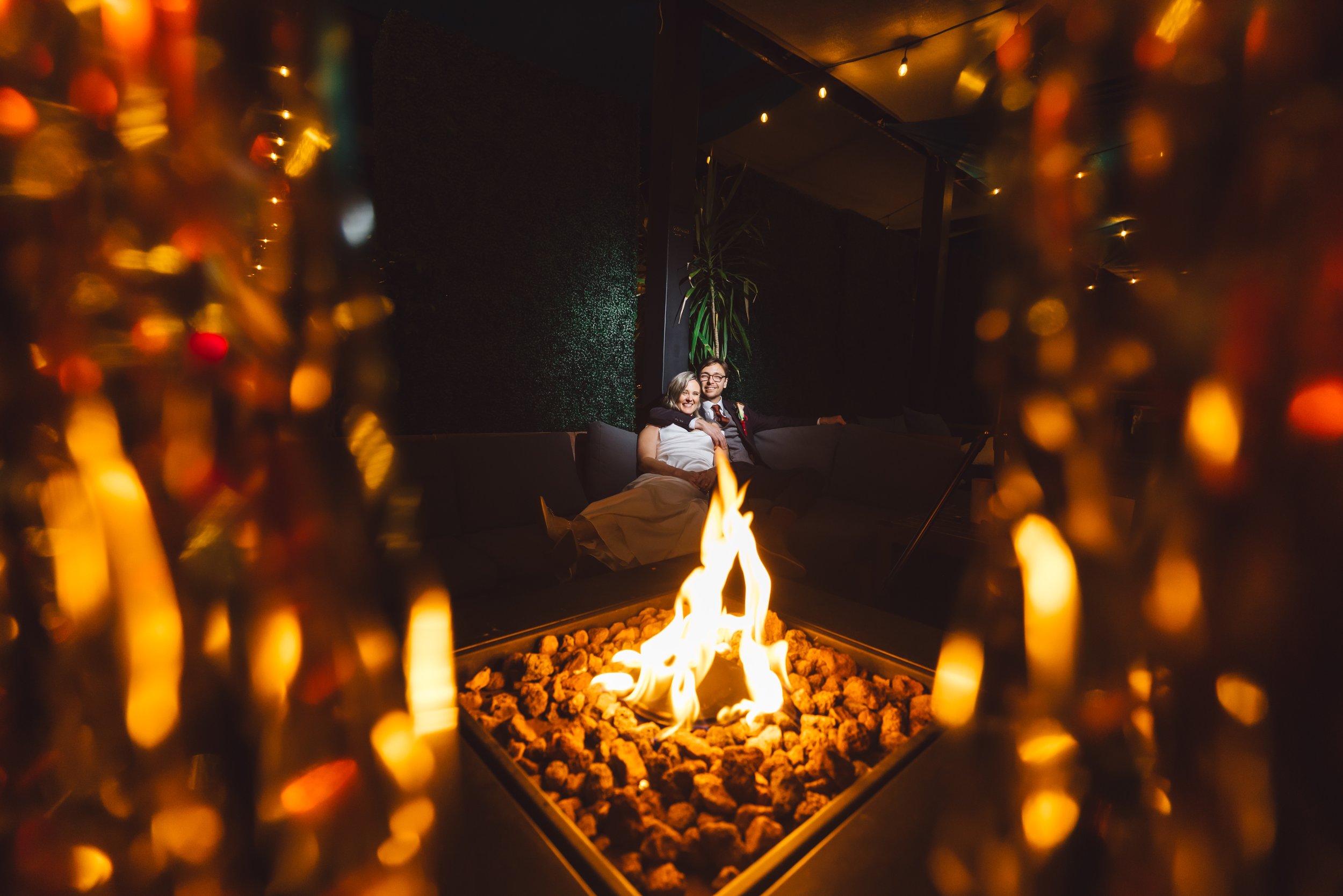 A couple sitting on a black lounge sofa in a dimly lit room, surrounded by a modern fire pit with rocks, seen through a decorative curtain, with string lights overhead and a tall plant in the background.