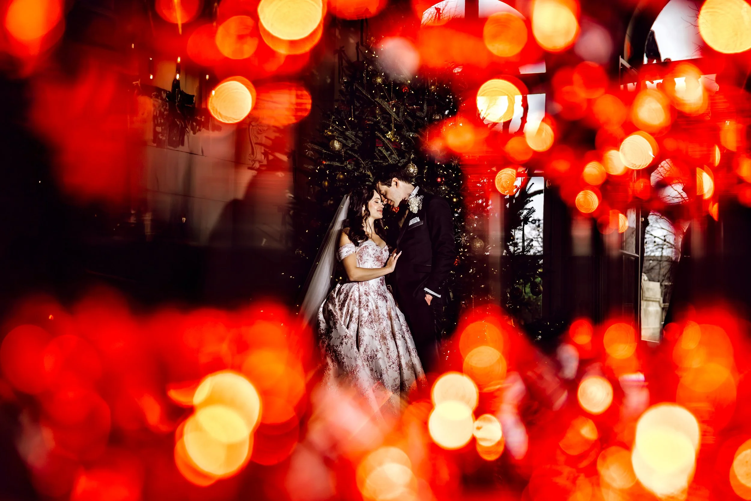 A bride and groom standing close together in front of a decorated Christmas tree, surrounded by warm red and orange bokeh lights.