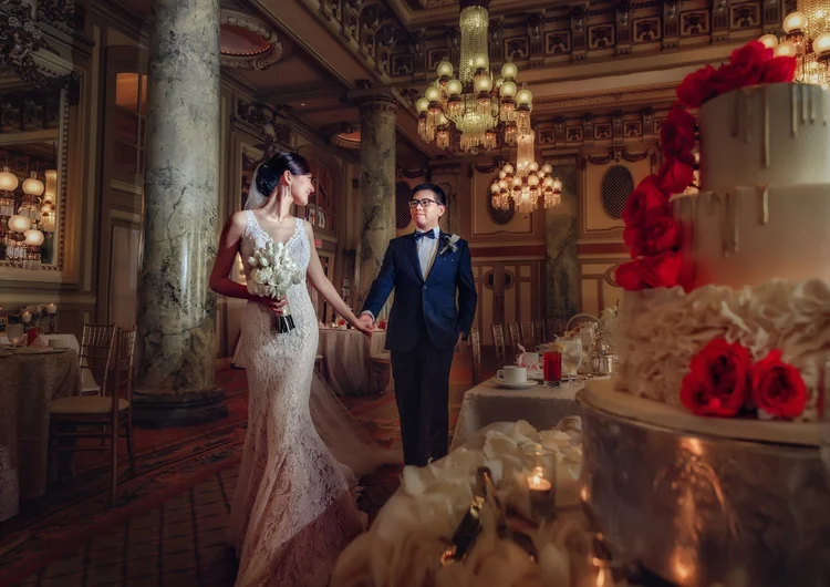 A bride and groom holding hands in a lavish wedding reception hall with ornate chandeliers, marble columns, and decorated tables. The bride is wearing a lace wedding gown and holding a bouquet, while the groom is in a tuxedo with a bow tie.