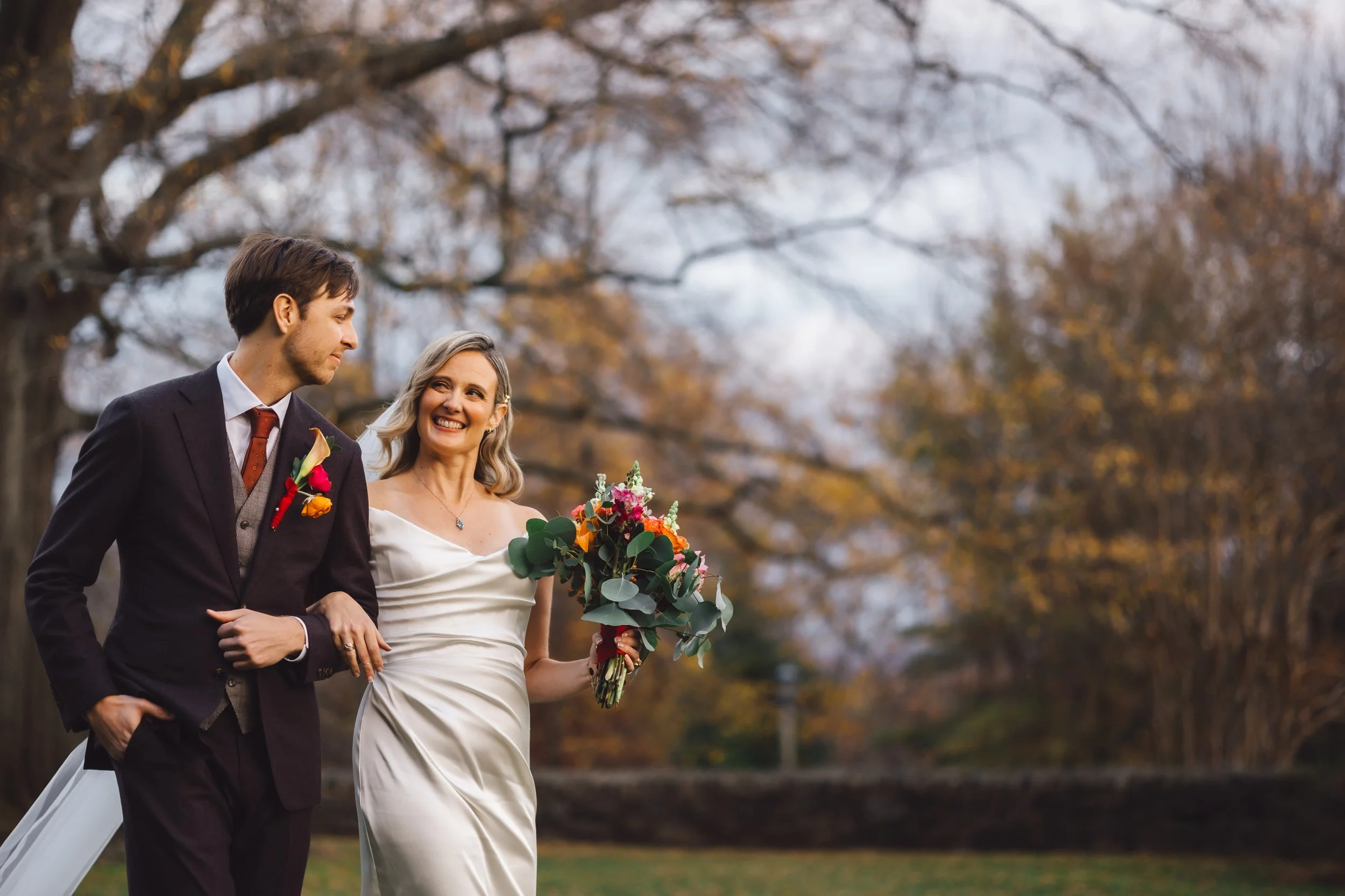 A bride and groom walking outdoors during fall, with the bride holding a bouquet of colorful flowers, as they smile at each other.