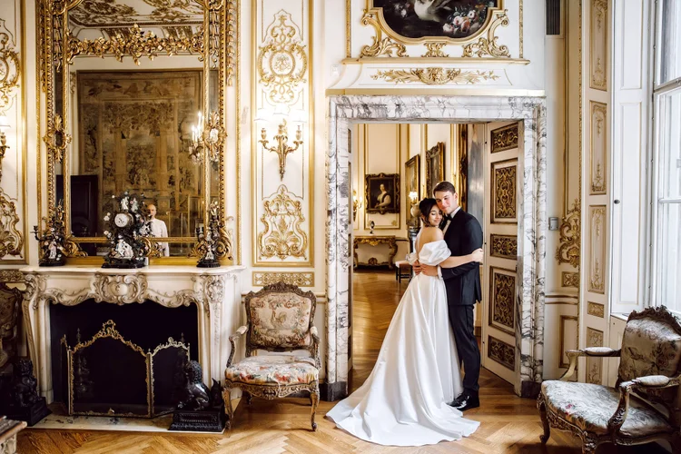 A bride and groom in formal wedding attire embracing in an ornate, luxurious room with gold decor and antique furniture, looking at the camera.
