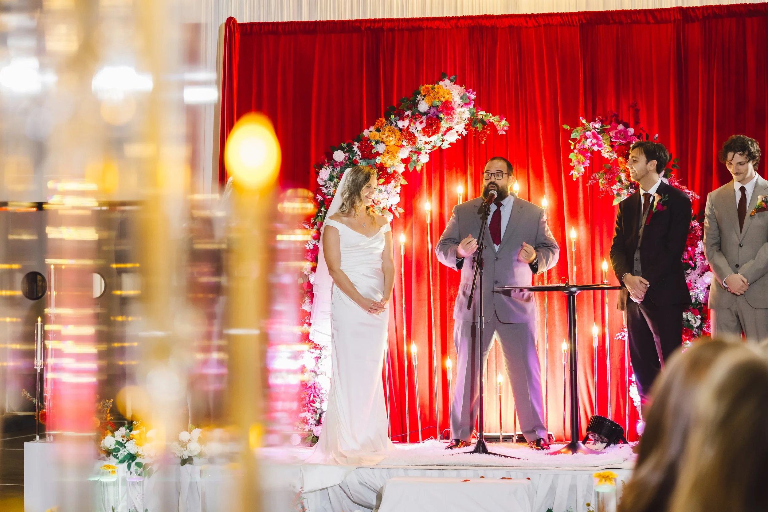 A wedding ceremony with the bride, groom, and officiant standing on a decorated stage with red curtains and floral arrangements.