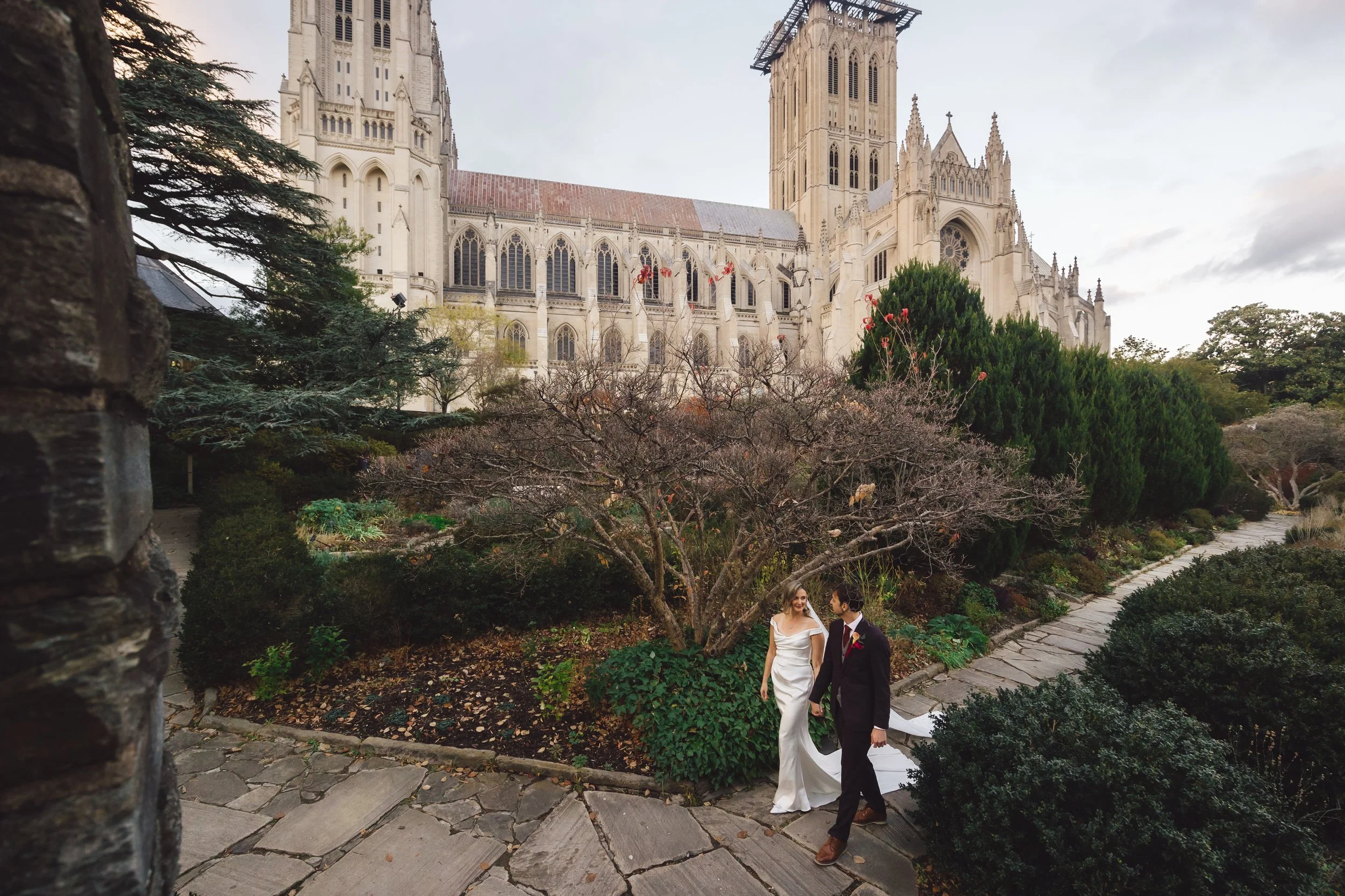A bride and groom holding hands and walking on a garden pathway near a large tree and bushes, with a historic Gothic-style cathedral visible in the background.