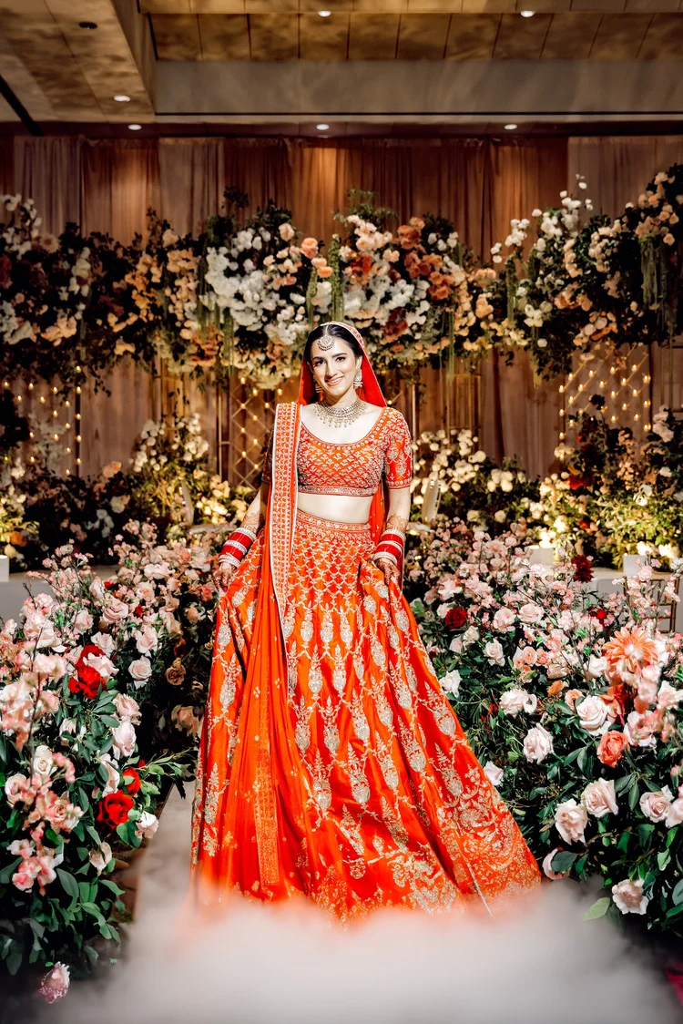 A woman in a traditional red and gold Indian bridal outfit standing amidst a decorated floral setting with pink and white flowers and fairy lights, smiling at the camera.