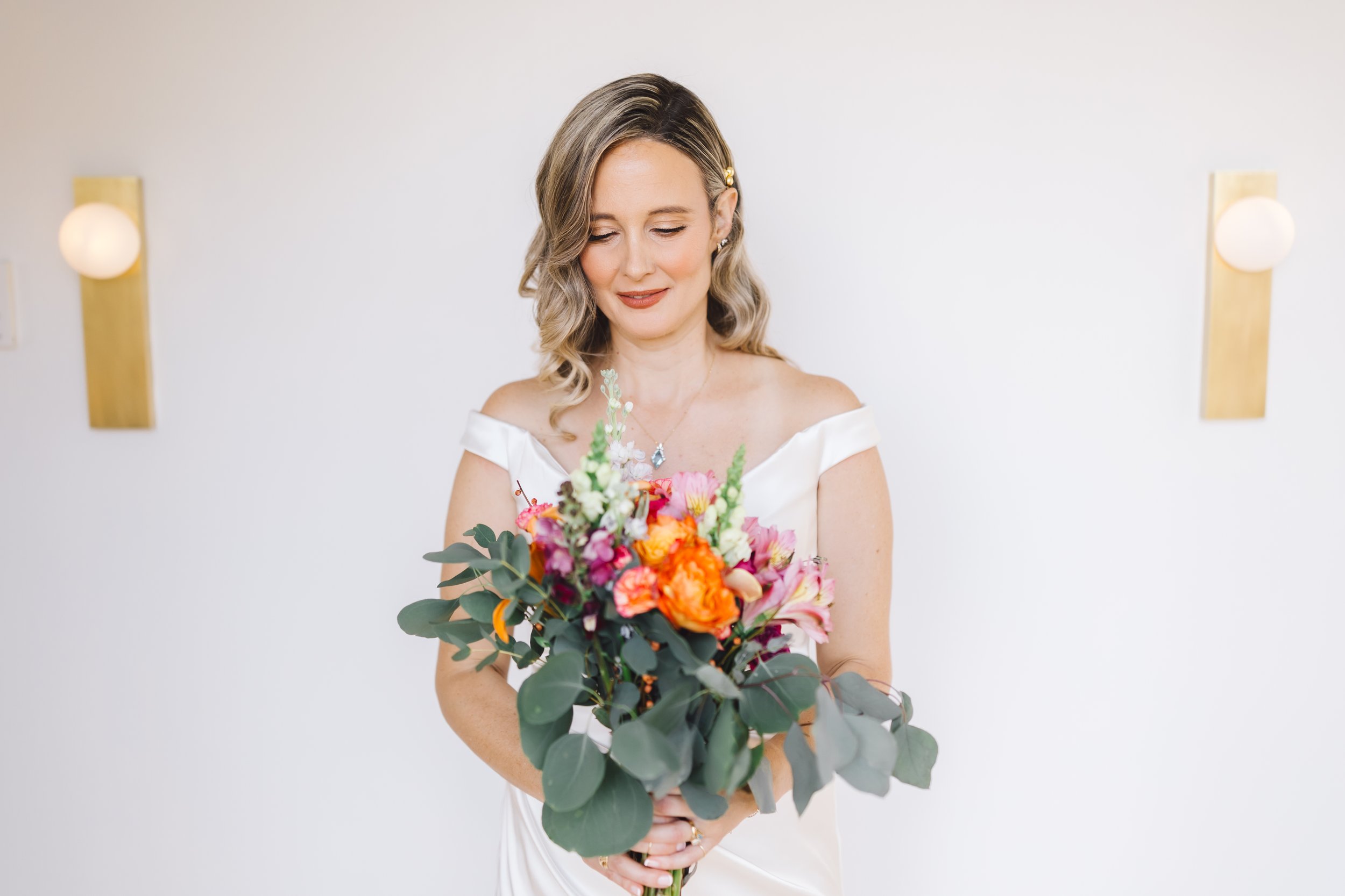 A woman in a white off-the-shoulder dress holding a colorful bouquet of flowers, standing in front of a plain white wall with two wall sconces.