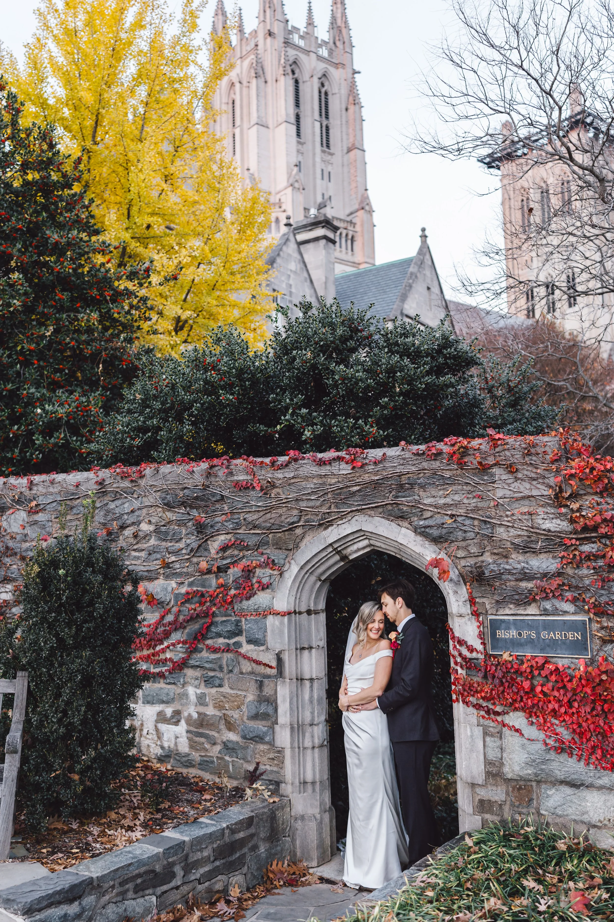 Wedding couple standing in a stone archway in Bishop's Garden, with a gothic church in the background, surrounded by autumn foliage.