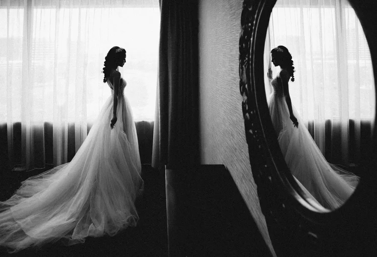 A black and white photo of a woman in a wedding dress standing in front of a window with sheer curtains, her reflection seen in a mirror.