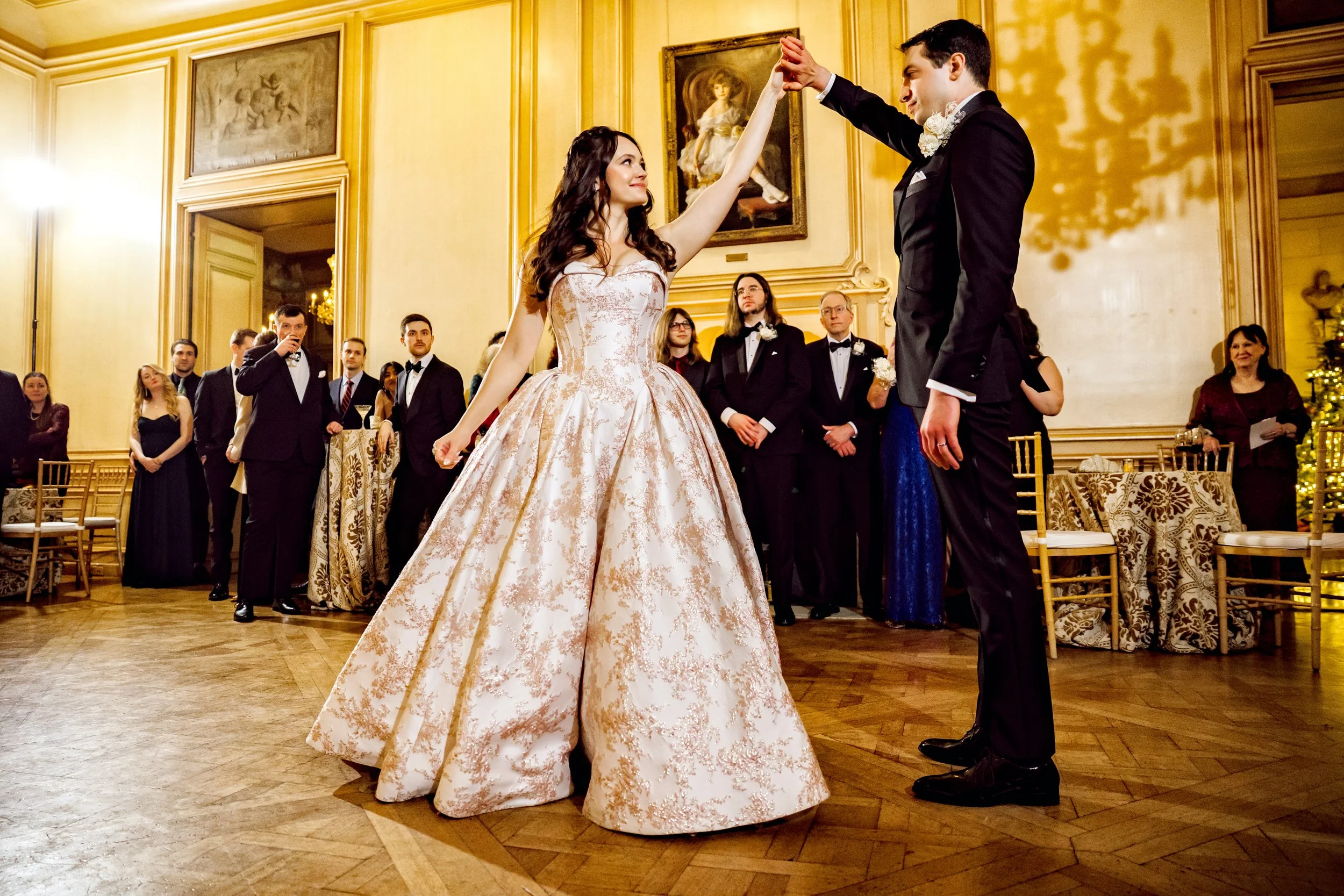 A bride and groom are dancing during their wedding reception, surrounded by guests in formal attire in an ornate hall.