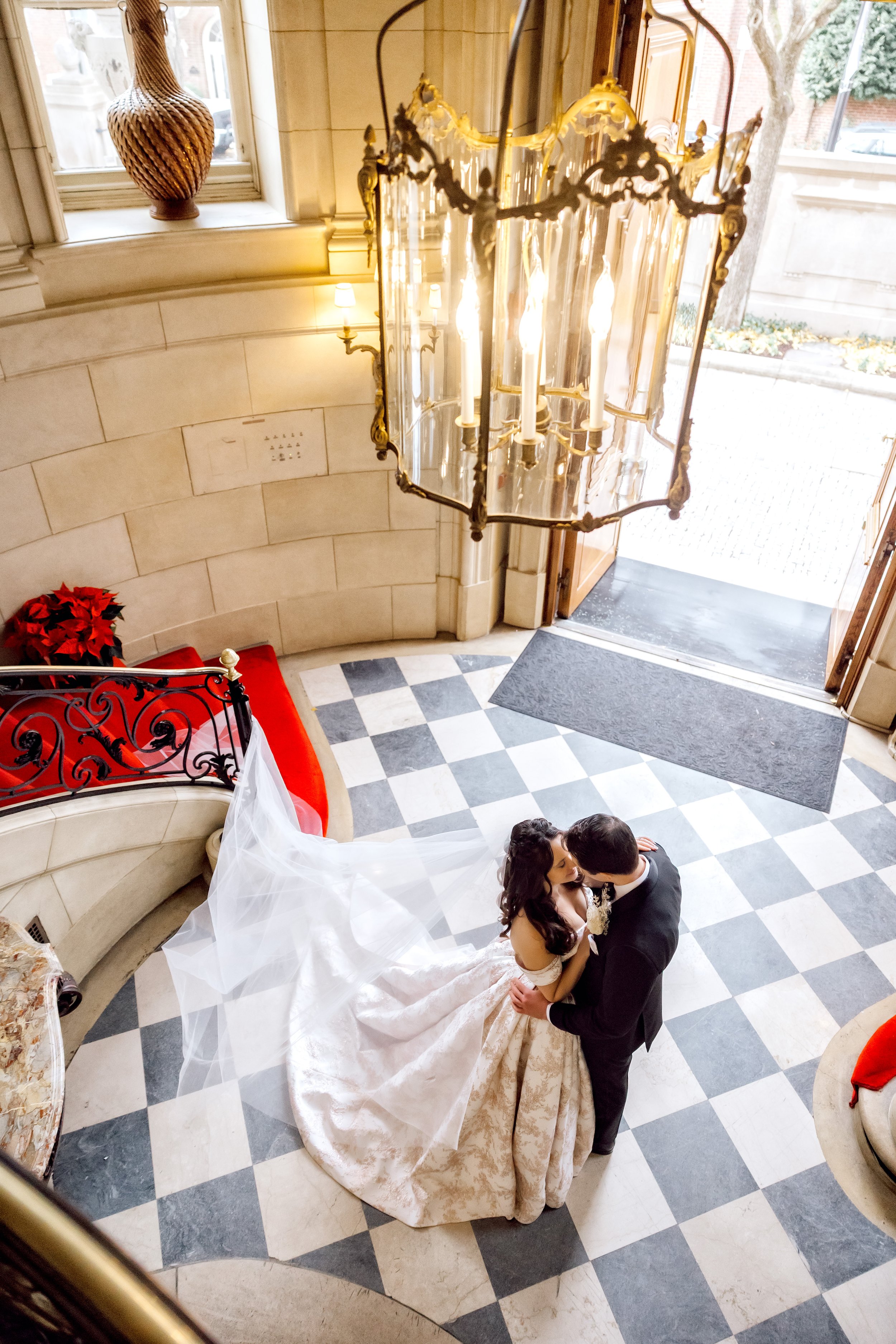 A bride and groom share a dance in the entrance hall of a building, seen from above. The bride is wearing a white wedding dress with a long train and veil, and the groom is dressed in a black tuxedo. The hall features checkered black and white floori