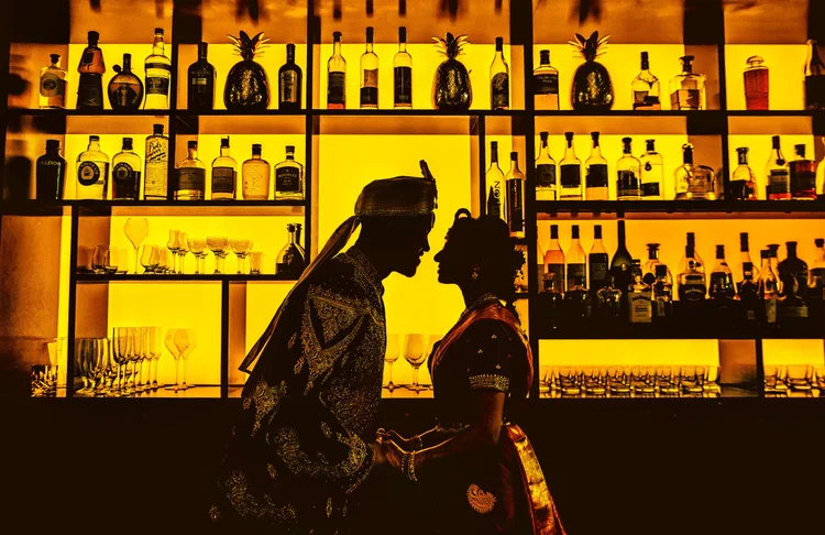 Silhouette of a couple in traditional attire holding hands in front of illuminated bar shelves filled with bottles and glassware.