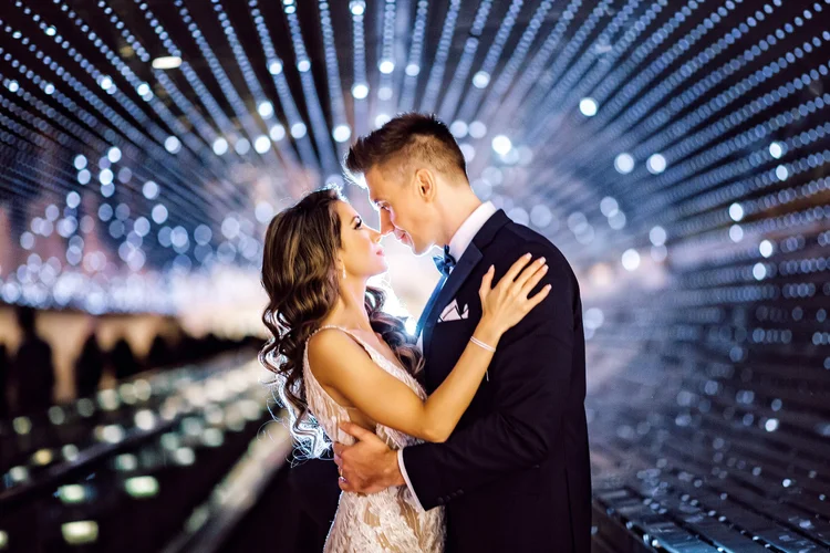 A couple in formal attire, about to kiss, standing close together in front of a background of decorative string lights.