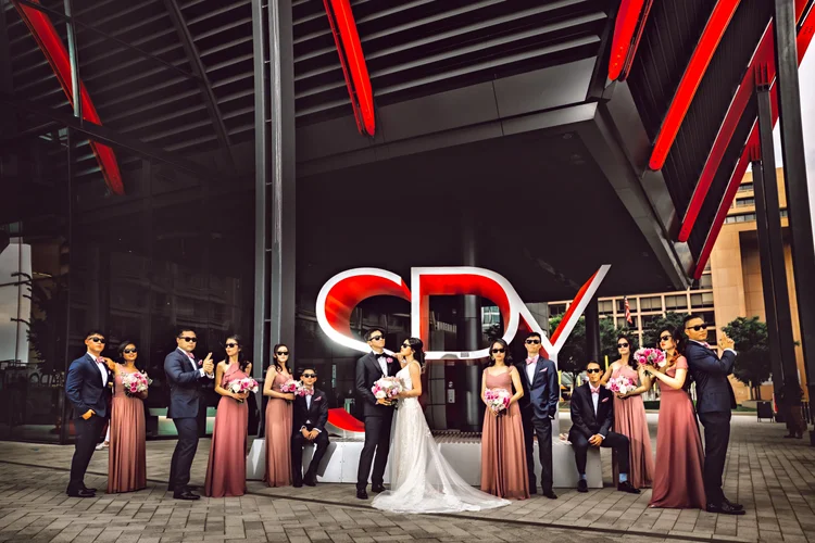 A wedding party posing in front of a modern building with a large red and white logo. The bride and groom are in the center, surrounded by bridesmaids and groomsmen, all dressed formally. The bride is wearing a white gown, and the bridesmaids are in 