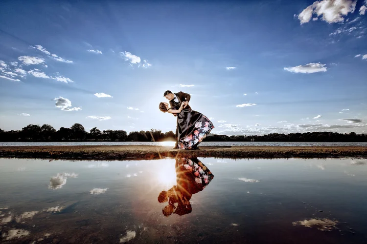 A couple dancing outdoors near a body of water at sunset, with their reflection visible in the water.