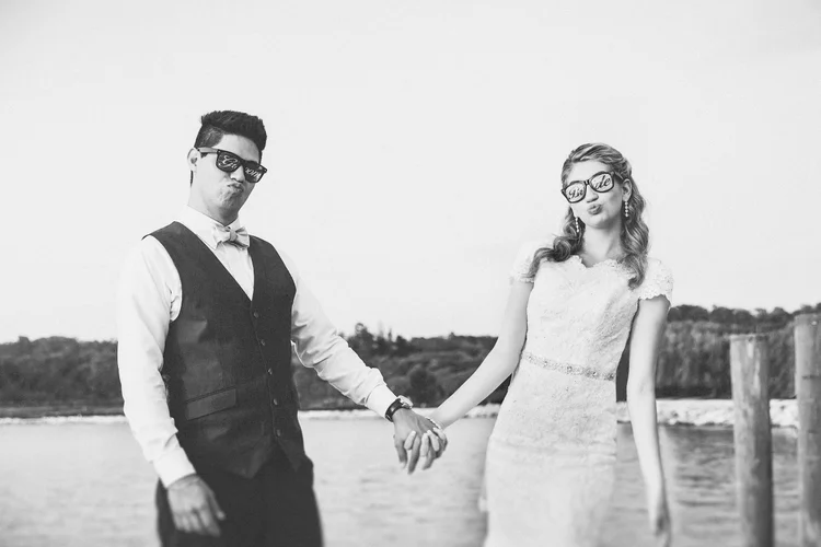 Black and white photograph of a couple holding hands on a beach, both wearing sunglasses with the words 'Love' and 'Bride,' the man in a vest and bowtie, the woman in a lace dress, with a distant shoreline in the background.