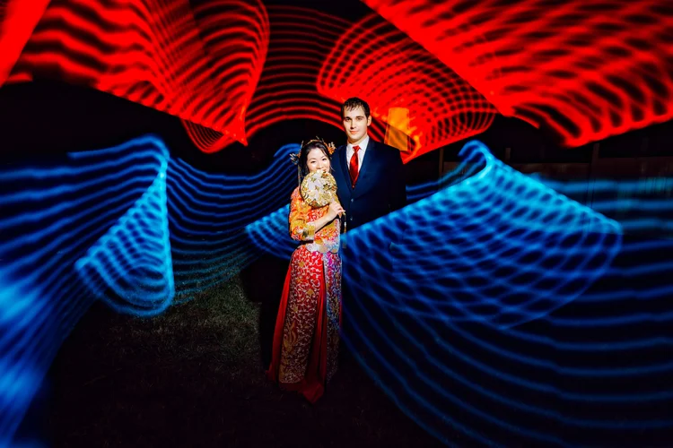 A couple dressed in formal attire standing under colorful light trails, with the woman holding a traditional hand fan.
