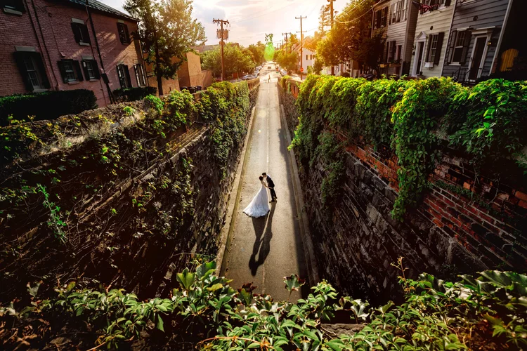 A bride and groom sharing a kiss in the middle of a narrow alley with high brick walls, greenery, and buildings on each side, during sunset.