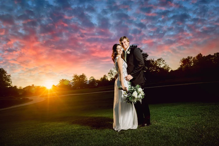 A bride and groom standing close together outdoors during sunset, with the bride holding a bouquet of white flowers and both smiling.