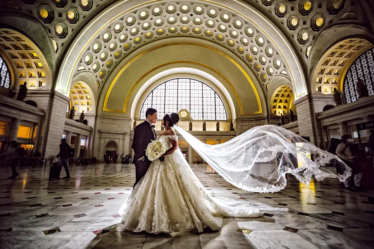 A bride and groom standing face to face inside a grand train station with a domed ceiling. The bride is holding a bouquet and wearing a wedding gown with a long veil flowing behind her. The groom is dressed in a tuxedo. There are a few people in the 