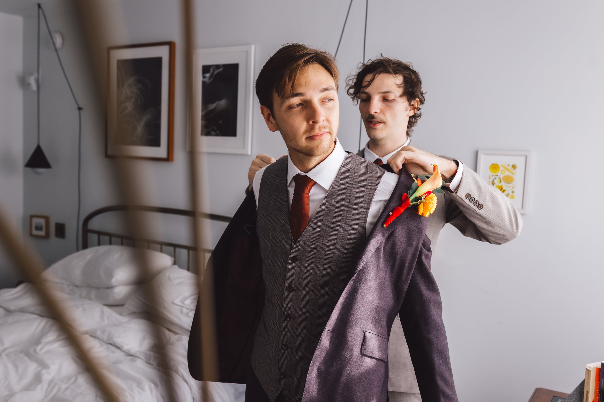 Two men in suits, one helping the other put on a jacket, in a bedroom with framed artwork on the wall.