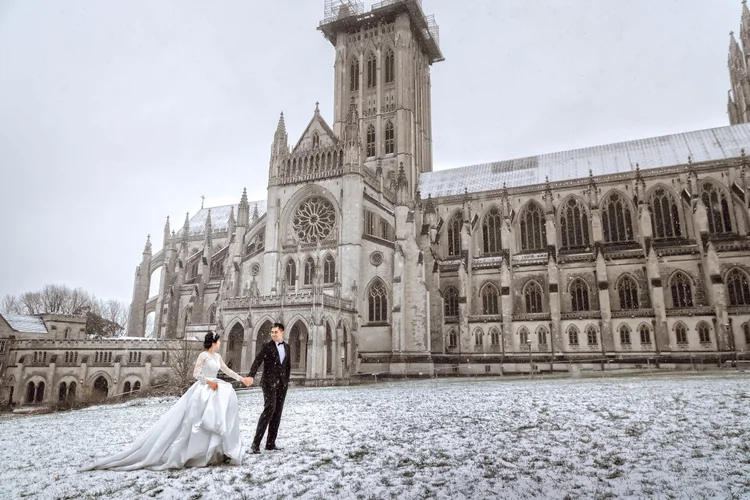 A bride in a white wedding gown and a groom in a black tuxedo hold hands outside a snowy gothic cathedral.