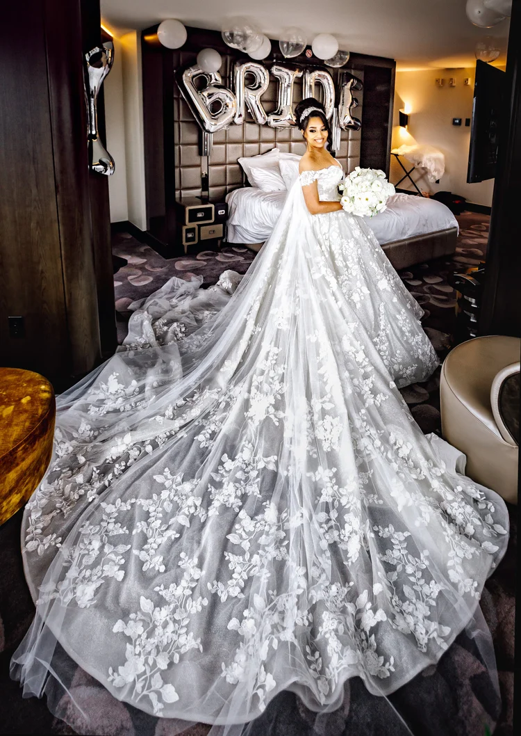 Bride in a wedding dress with a long, lace-embellished train, smiling and holding a bouquet of white flowers in a hotel room decorated with silver balloons spelling 'BRIDE' in the background.