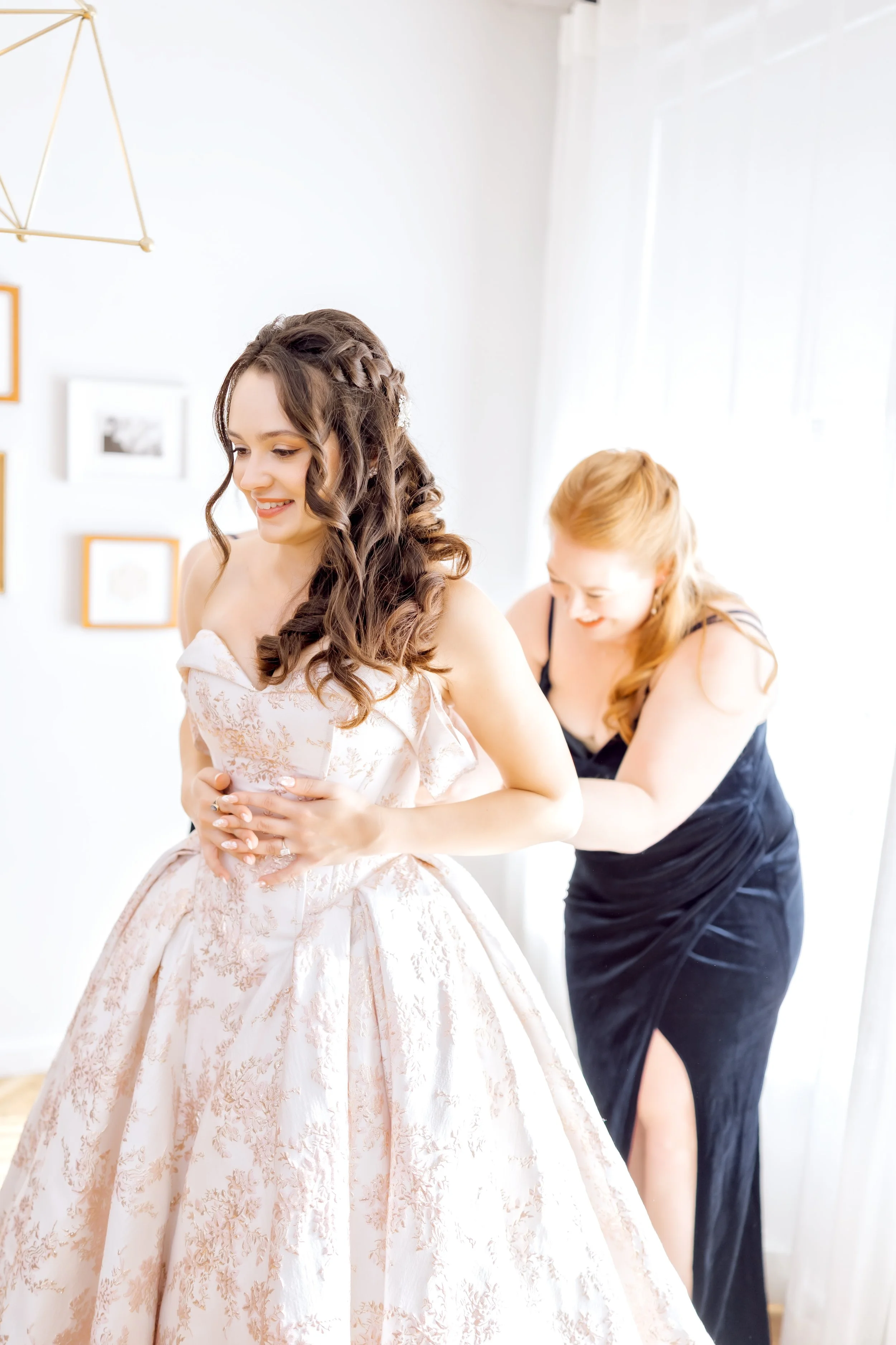 A woman in a pink floral wedding dress getting help from a woman in a black dress with a slit on her thigh to zip up or adjust the dress, in a brightly lit room with white walls and framed pictures.