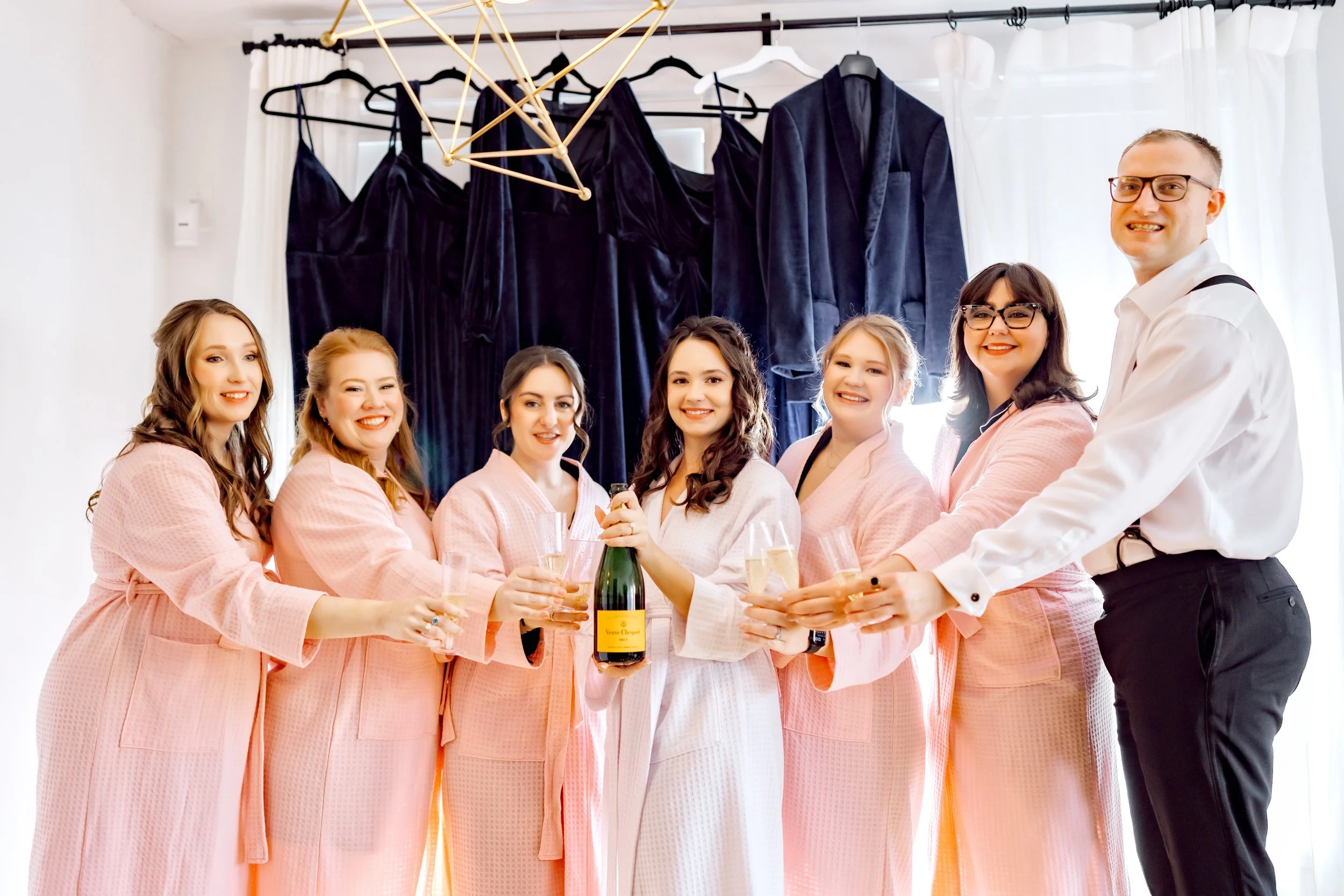 Group of bridesmaids and a groom celebrating with champagne before a wedding, with black dresses hanging in the background.