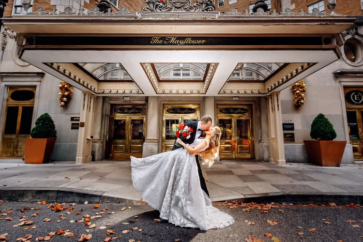 A bride and groom kissing in front of a building with the sign 'The Mayflower', the groom holding a bouquet of red roses, as fallen leaves are scattered on the street.